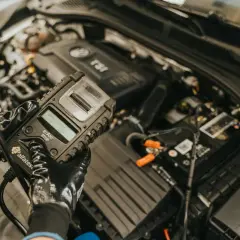 Mechanik w rękawiczkach sprawdza stan akumulatora za pomocą testera, który może również diagnozować czujnik halla w samochodzie.