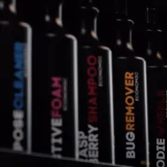 Rząd czarnych butelek z etykietami: "POSECLEANER", "CTIVEFOAM", "RASPBERRY SHAMPOO", "BUGREMOVER", "ODIE WHEELS", "ASSCLEANER". To są kosmetyki do samochodu.