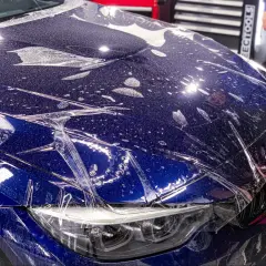 Nakładanie folii ochronnej na maskę ciemnoniebieskiego BMW. Widać krople wody i pęcherzyki powietrza pod folią.