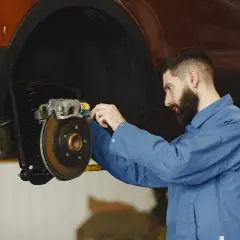 Mechanik w niebieskim kombinezonie pracuje przy hamulcach samochodu. Widać tarczę hamulcową i zacisk. To ważny punkt, od dot 3 a dot 4 zależy bezpieczeństwo jazdy.