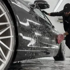 Mężczyzna w czapce i okularach myje czarne auto gąbką. Widać pianę i felgi. Dowiedz się, czym umyć auto domowe sposoby.