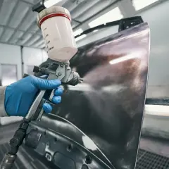 Ręka w niebieskiej rękawiczce trzyma pistolet lakierniczy, nanosząc warstwę na element karoserii. Czy na podkład epoksydowy można kłaść lakier? Tak, to kluczowy etap renowacji.