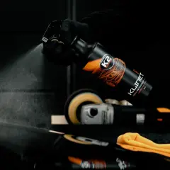 Rękawiczka w czarnej rękawiczce rozpyla produkt K2 KLINET na czarny lakier samochodowy. W tle polerka.