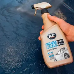 Dłoń trzyma butelkę z woskiem w sprayu K2 SPID WAX, gotową do usunięcia mlecznych plam na lakierze samochodowym.