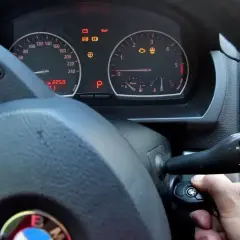 Ręka próbuje odpalić auto, ale kontrolki świecą. Kluczyk w stacyjce, a na desce rozdzielczej widoczne m.in. kontrolka silnika i ABS.