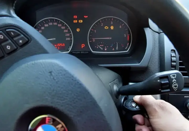 Ręka próbuje odpalić auto, ale kontrolki świecą. Kluczyk w stacyjce, a na desce rozdzielczej widoczne m.in. kontrolka silnika i ABS.