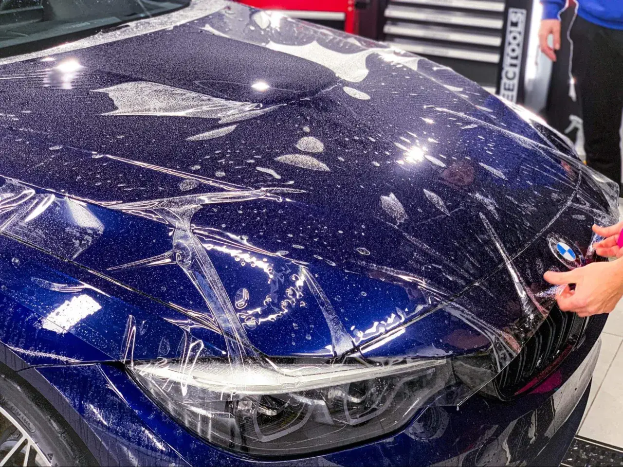 Nakładanie folii ochronnej na maskę ciemnoniebieskiego BMW. Widać krople wody i pęcherzyki powietrza pod folią.