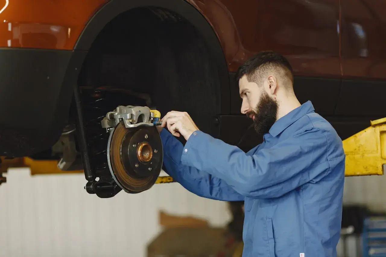 Mechanik w niebieskim kombinezonie pracuje przy hamulcach samochodu. Widać tarczę hamulcową i zacisk. To ważny punkt, od dot 3 a dot 4 zależy bezpieczeństwo jazdy.