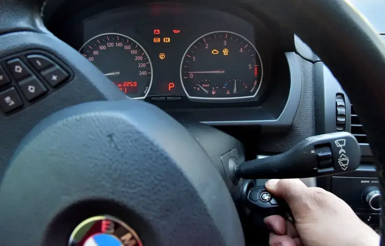 Ręka próbuje odpalić auto, ale kontrolki świecą. Kluczyk w stacyjce, a na desce rozdzielczej widoczne m.in. kontrolka silnika i ABS.