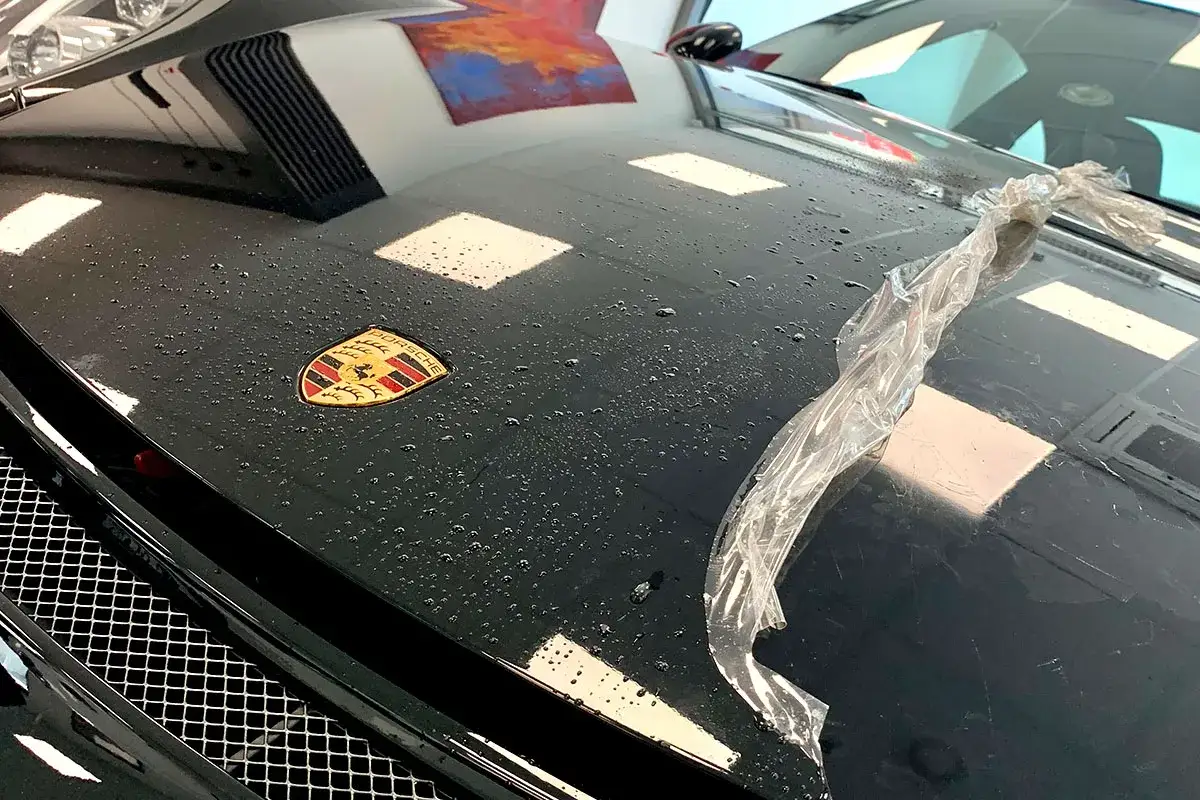 Czarna maska Porsche z logo, pokryta kropelkami wody. Widać fragment folii PPF, która chroni lakier.