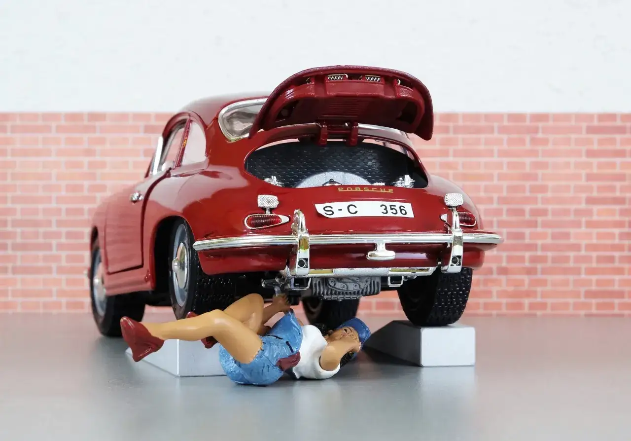 Miniaturka Porshe 356 na podwyższeniu, z otwartą klapą silnika. Pod samochodem leży figurka kobiety, która wykonuje wymianę oleju na górce.